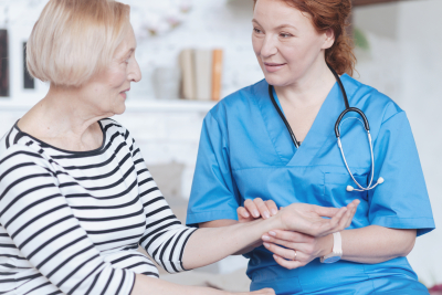 Selective focus on a female caregiver looking at a senior lady while sitting next to her on a sofa and measuring her pulse at home.